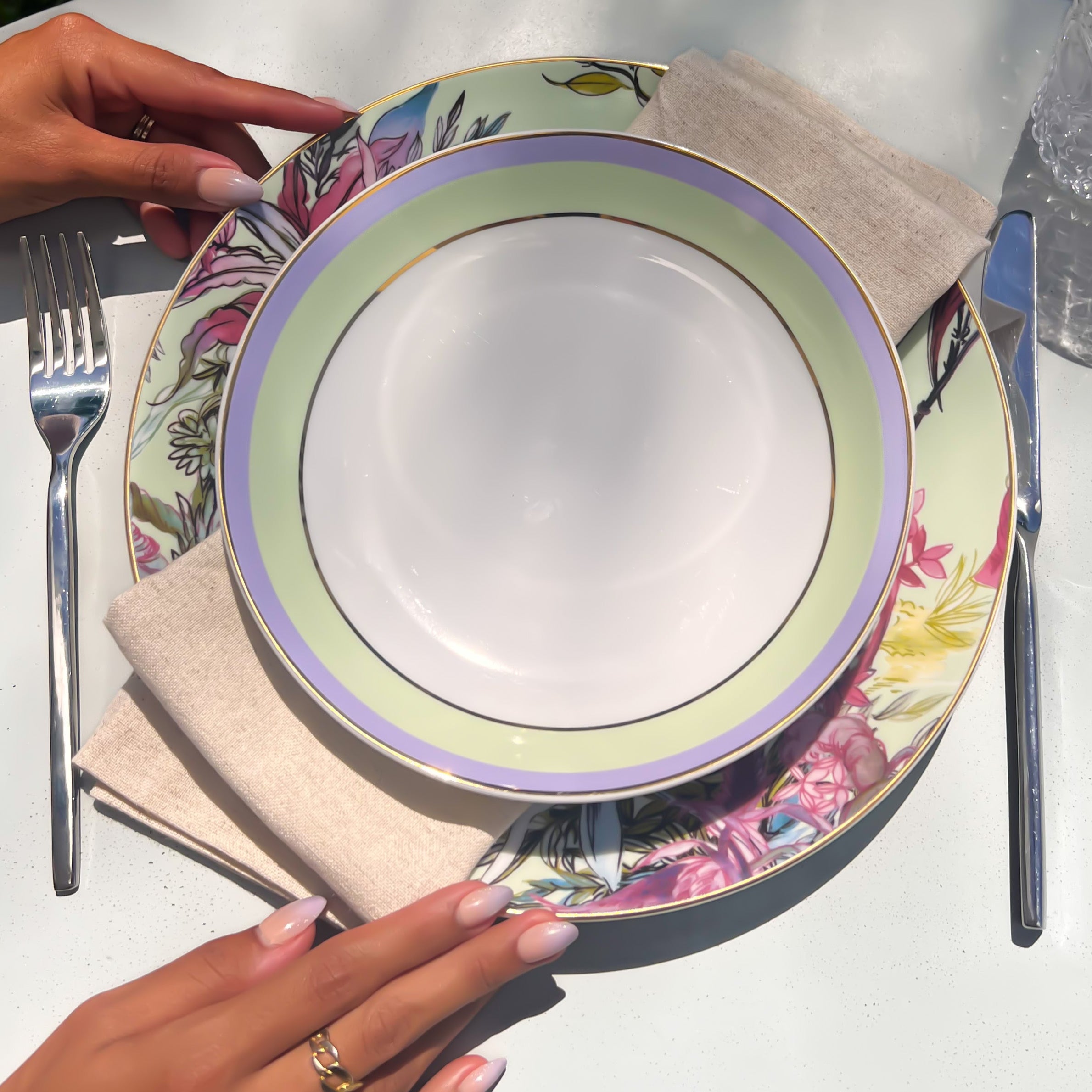 Person setting a table with floral plates, silverware, and a glass on a grassy background.