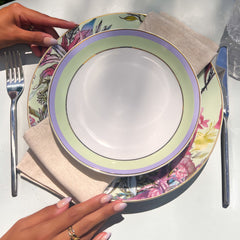 Person setting a table with floral plates, silverware, and a glass on a grassy background.